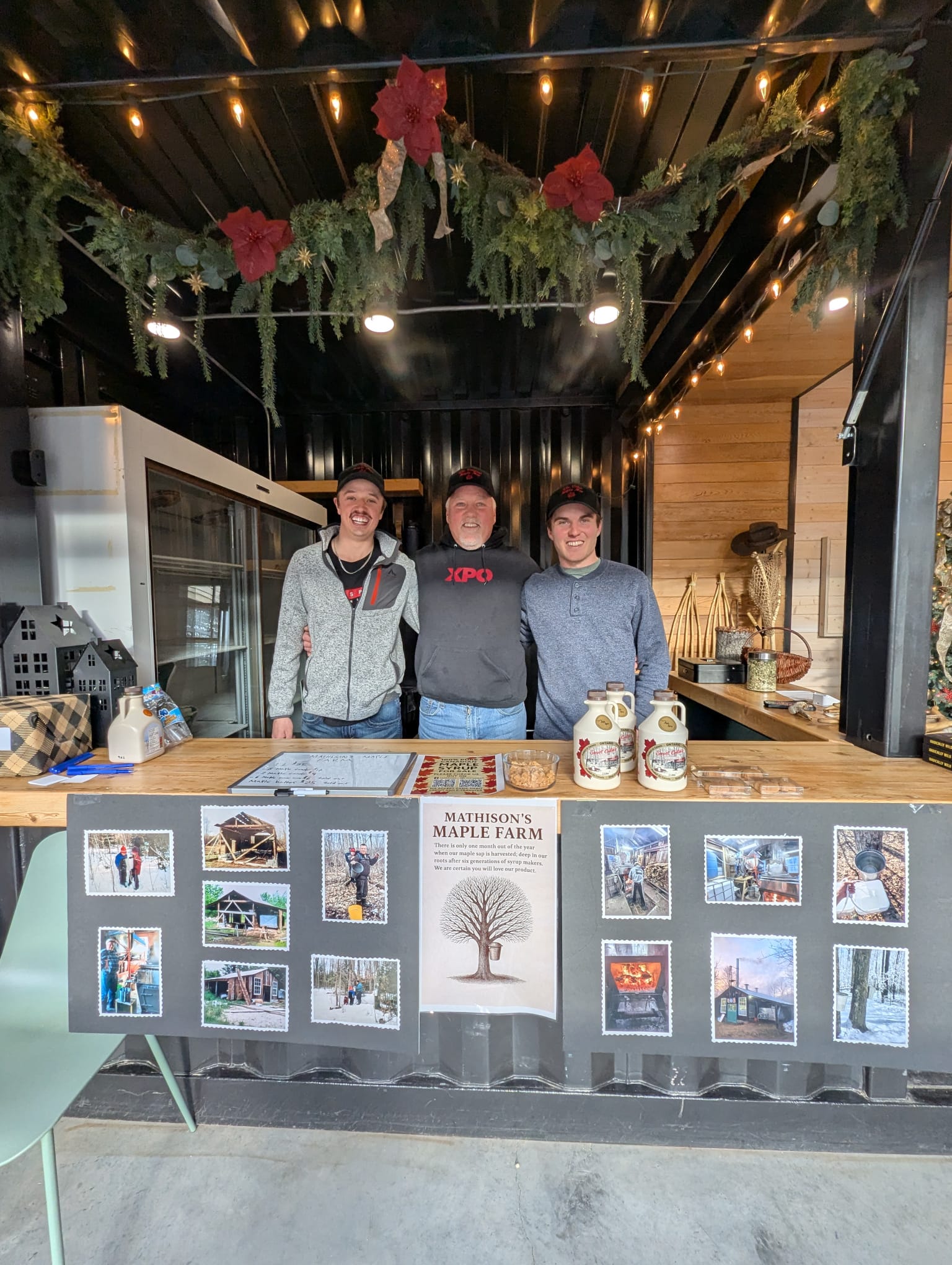 Three people standing behind a counter with decorative elements and a display of maple syrup.